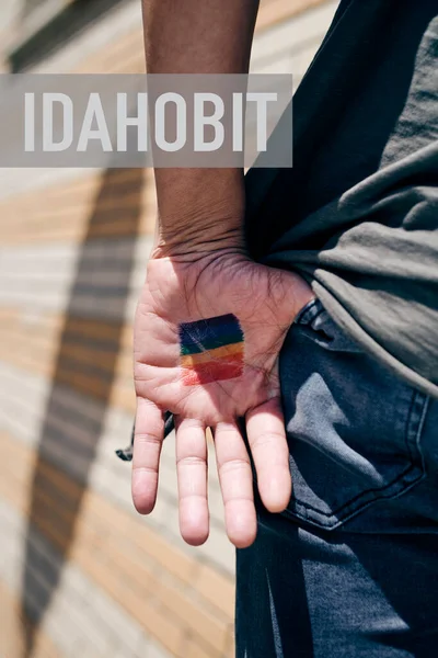 Genç bir insanın avucundaki gökkuşağı bayrağına yakından bakın, arkadan görünüyor, ve IDAHOBIT metni, Uluslararası Homofobi, Bifobi, İnterfobi ve Transfobi Karşıtı Günler için.