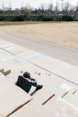 genç bir beyaz adam, arkadan görülen, kot pantolon ve gri kapüşonlu ceket giyen, bir açık beton merdiven üzerinde oturan kıvrılmış
