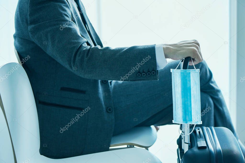 closeup of a young businessman, in a gray suit, sitting in the waiting hall of an airport or a train station, holding a surgical mask while is leaning his hand on the telescopic handle of his suitcase