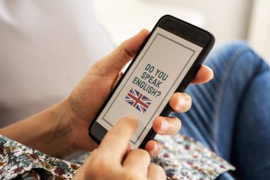 Kafkasyalı genç bir adamın resmi, günlük kıyafetler giymiş, kapalı bir koltukta oturuyor, akıllı telefonu elinde metinle ekranında İngiliz bayrağı ve İngilizce konuşuyor musunuz?
