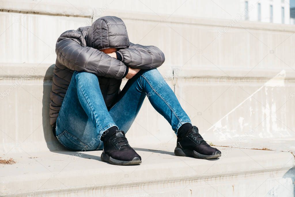 a young caucasian man, wearing jeans and a gray hooded jacket, curled up sitting on an outdoor concrete stairway