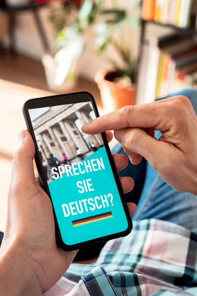 カジュアルな服を着た若い白人男性が椅子に座ってスマートフォンを手にテキストを持っているドイツ語で書かれたドイツ語をスクリーンで話す — ストック写真