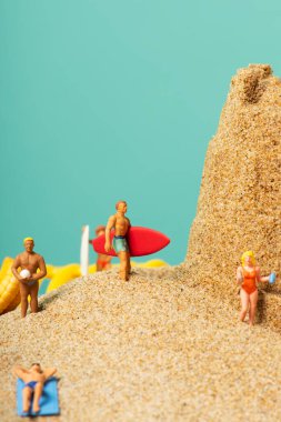 Mayolu minyatür insanlar sörf tahtaları taşıyorlar ya da kumsalın kumsalında, kumdan kalenin yanında, üzerinde boş bir alan olan mavi bir zemine yaslanıyorlar.