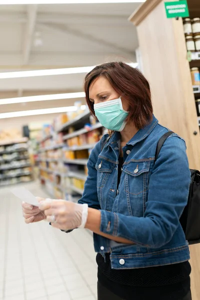 Maskeli ve listeli beyaz kadın süpermarkette görünüyor.
