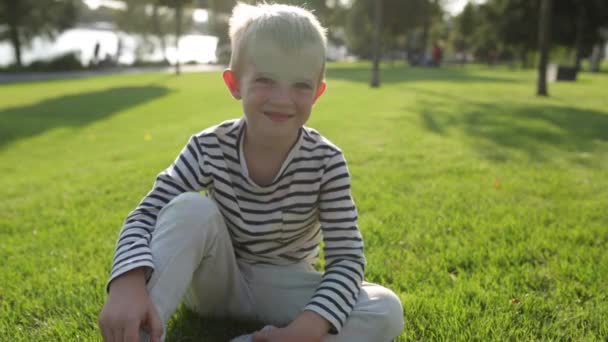 Χαριτωμένο όμορφο ευτυχισμένο μικρό αγόρι κουνώντας το κεφάλι, ενώ κάθεται στο γρασίδι — Αρχείο Βίντεο