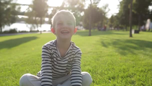Χαριτωμένο όμορφο ευτυχισμένο μικρό αγόρι χαμογελά, κάθεται στο γρασίδι — Αρχείο Βίντεο