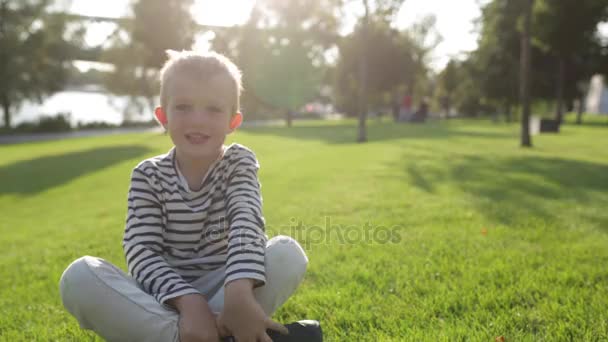 Χαριτωμένο όμορφο ευτυχισμένο μικρό αγόρι παίζει με στόμα, κάθεται στο γρασίδι — Αρχείο Βίντεο