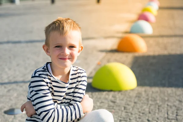 Søt, vakker, liten rødhåret gutt som smiler og sitter på fargede steiner – stockfoto