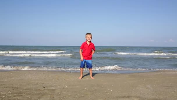 Fröhlicher kleiner aufgeregter aktiver Junge springt am Strand in Meeresnähe — Stockvideo