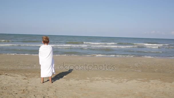 Criança bonita feliz em toalha branca olhar para longe das ondas do mar, vento na praia — Vídeo de Stock