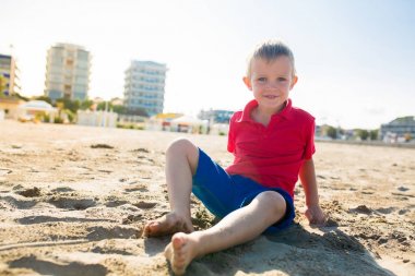 Mutlu güzel gülümseyen bir çocuk oturuyor kum plaj, bak kameraya