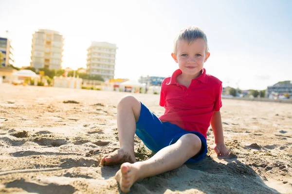 Feliz hermoso niño sonriente sentado en la playa de arena, mira a la cámara — Foto de Stock