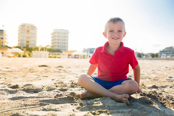 Feliz hermoso niño sonriente sentado en la playa de arena, mirando a la cámara — Foto de Stock