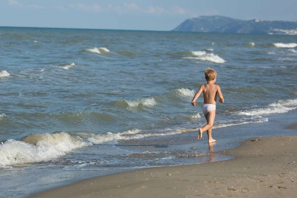 Glad liten pojke som kör långt bort längs en strand vid havet Stockbild