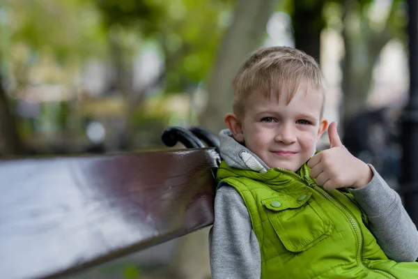 Gelukkig blond jongetje Toon ok teken zittend op een bankje, camera kijken — Stockfoto