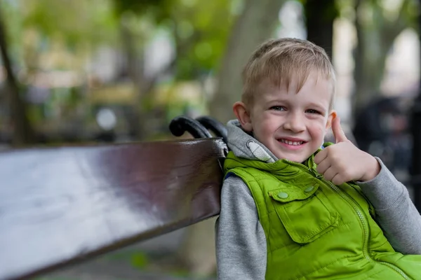 Feliz niño rubio sonriente mostrar ok signo sentado en un banco, mirando a la cámara —  Fotos de Stock