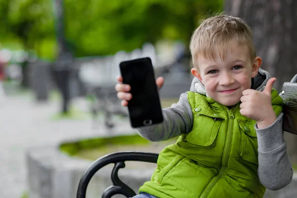 Criança feliz mostrando celular, telefone inteligente, sentado em um banco da cidade — Fotografia de Stock