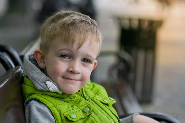 Ευτυχής χαμογελαστοί μικρό ξανθό αγόρι κάθεται σε ένα παγκάκι που βλέπουν τα φωτογραφικών μηχανών Royalty Free Φωτογραφίες Αρχείου