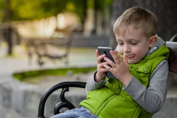 Feliz criança interessada jogar celular, telefone inteligente, sentado em um banco da cidade — Fotografia de Stock