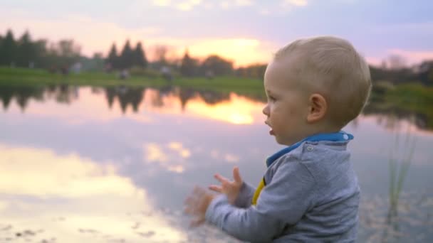 Un niño pequeño se sienta en el lago y observa el atardecer — Vídeos de Stock