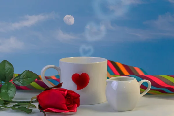 White mug and red heart with red rose, on blue sky background.