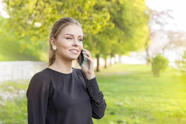 Χαρούμενος νεαρός καυκάσιος γυναίκα που μιλάτε στο τηλέφωνο, πράσινο φύλλα θολή φόντο. Άνοιξη — Φωτογραφία Αρχείου