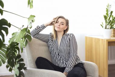 Sarışın gülümseyen ve çerçeve arıyorum. Çizgili bir gömlek kız büyük yeşil houseplant karşı bir sandalyede oturur