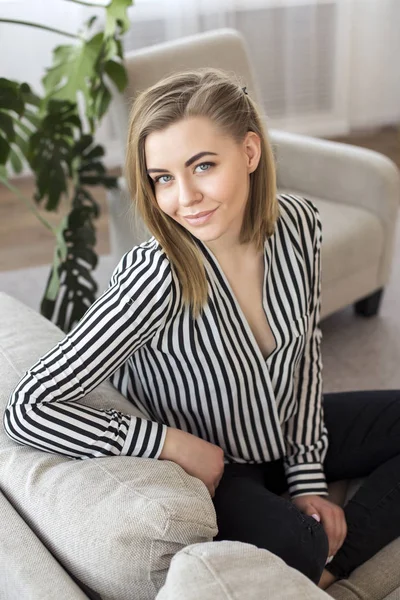 Attractive blonde in a striped blouse is sitting on the couch in her apartment — Stock Photo, Image