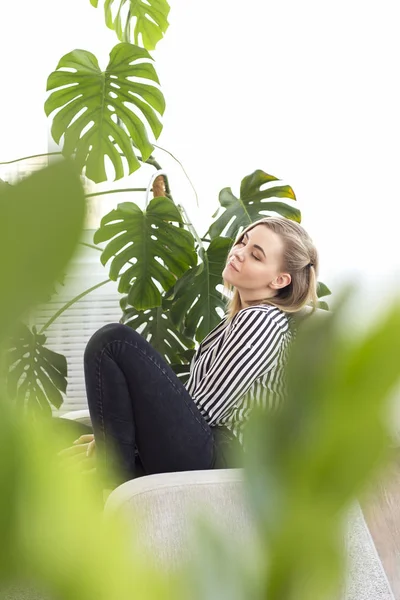 Jonge blonde rust thuis. Tegen de achtergrond van een grote kamerplant — Stockfoto