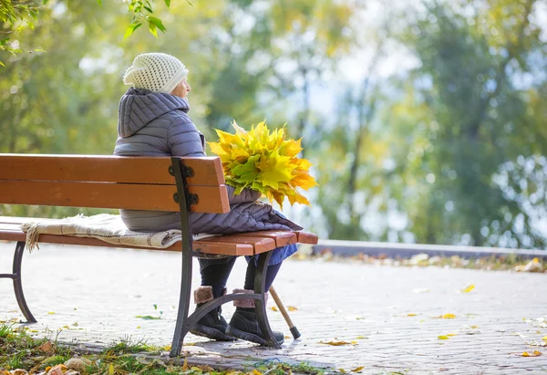 Senior žena sedí na lavičce v parku a drží spoustu podzimních listí — Stock fotografie