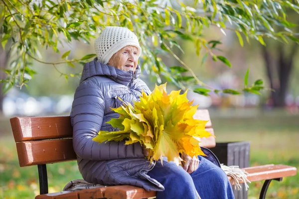 Mulher sênior sentada no banco no parque e segurando um monte de folhas de outono — Fotografia de Stock