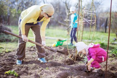 Küçük kızı arka planda oğlu ve oğluyla oynarken genç bir kadın bahçede çalışıyor.