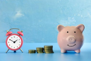 On a dark blue background photographed piggy bank, next to her a small pink alarm clock and three stacks of coins. Preservation of savings. clipart