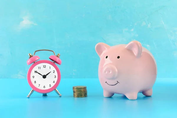 背景色が水色にピンクの貯金。横にコインと小さなピンクの目覚まし時計のスタック。明るい画像. — ストック写真