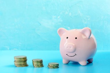 A pink pig piggy bank on a blue background. Nearby is a three stacks of coins. clipart