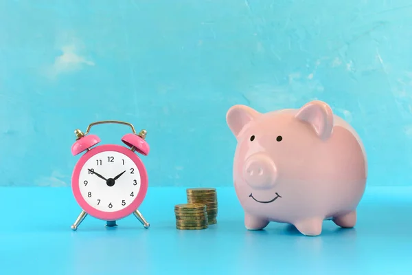 Piggy bank on a turquoise background, next to a small pink alarm clock and two stacks of coins.