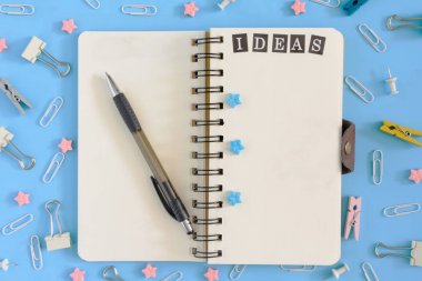 Yaylar üzerinde not defteri açık yatıyor. Karışıklık büro malzemeleri yanında. Defterin beyaz sayfaları yazıt fikirler ve mavi yıldız içerir. Mavi bir arka plan üstüne fotoğraf.