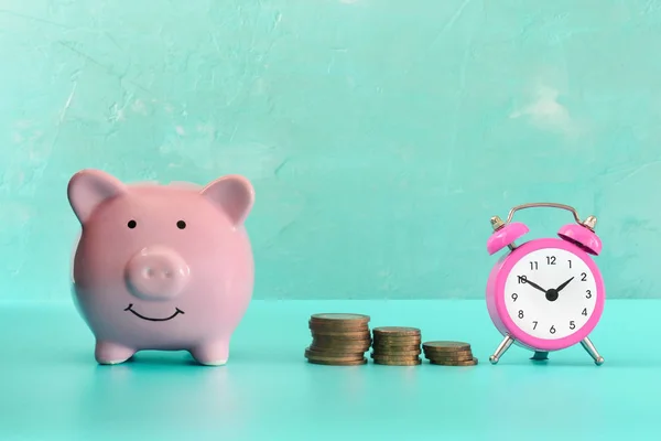 Entre o porquinho e um pequeno despertador rosa, três pilhas de moedas estão em ordem. O símbolo do crescimento do dinheiro no tempo. Negócios ainda vida em fundo brilhantemente turquesa . — Fotografia de Stock