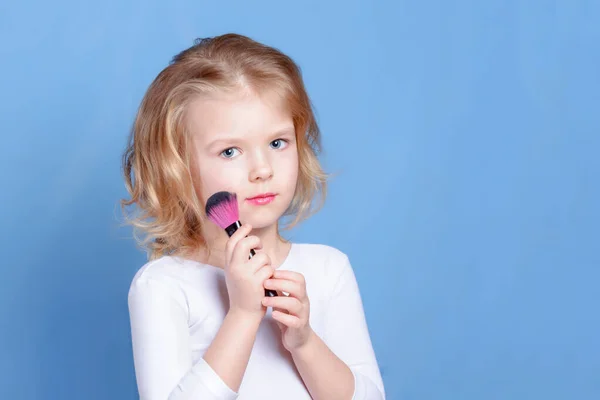 Una hermosa niña sostiene un cepillo de maquillaje en su mano. Una niña pequeña con el pelo rubio. Luz publicidad foto . — Foto de Stock