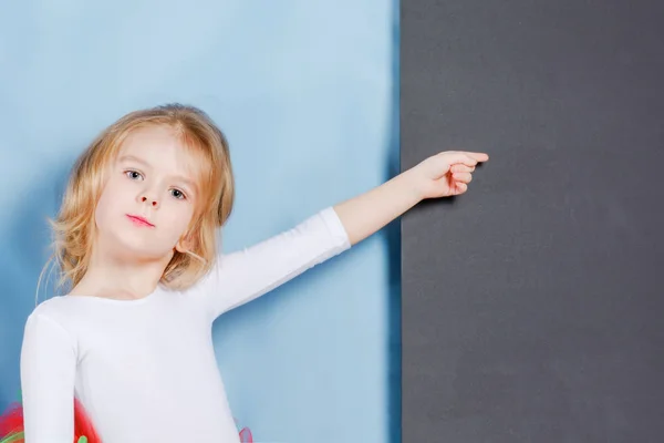 Das kleine Mädchen mit den blonden Haaren zeigt einen Finger auf leeren Raum auf schwarzem Hintergrund. schönes süßes Baby. — Stockfoto