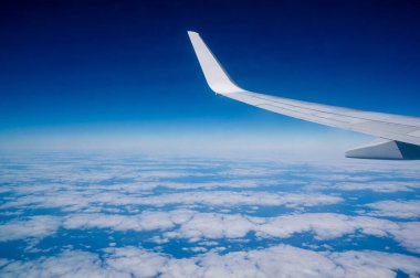 Yolcu penceresinden mavi gökyüzünün altında yüksek irtifada bulutların üzerinde uçan bir uçağın kanat görüntüsü