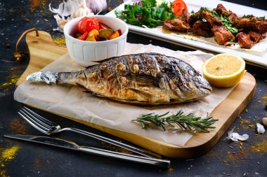 Yağlı kağıt üzerinde ızgara balık ve ahşap kesim tahtası. Etrafı ızgara domateslerle çevrili. Izgara balık dorada bbq hala hayatta.