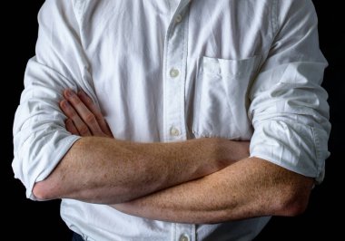 Beyaz gömlekli adam elleri göğsünde çaprazlanmış, koyu arkaplanda poz veriyor.