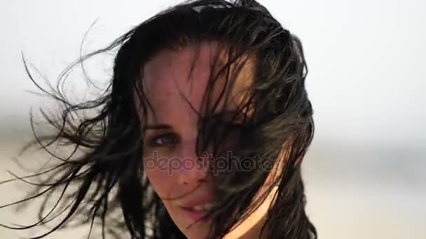 Retrato de una hermosa chica sonriendo en la playa al atardecer con el pelo ondulado . — Vídeos de Stock