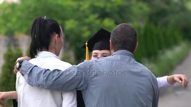 Outdoor scene of beautiful female graduating student dressed in cup and gown,hugging her parents. — Stock Video