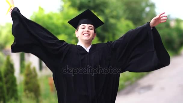 Outdoor scene of beautiful female graduating student dressed in cup and gown jumping up. — Stock Video