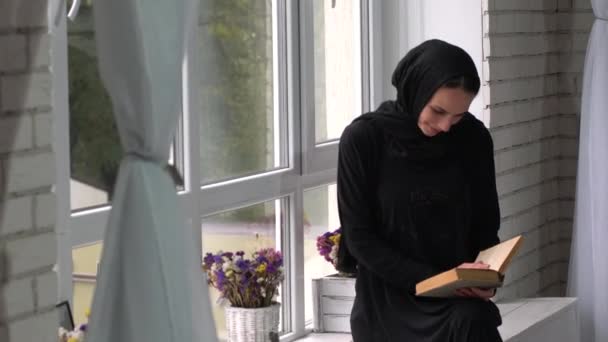 Hermosa entrometida joven oriental leyendo libro al lado de la ventana . — Vídeos de Stock