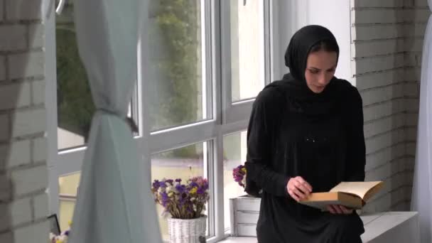 Beautiful mengen Oost-jonge vrouw lezen boek naast venster. — Stockvideo