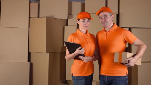 Correos femeninos anf masculinos en uniforme naranja de pie contra cajas de cartón marrón en el fondo. Personal de la empresa de entrega, disparo 4K — Vídeos de Stock