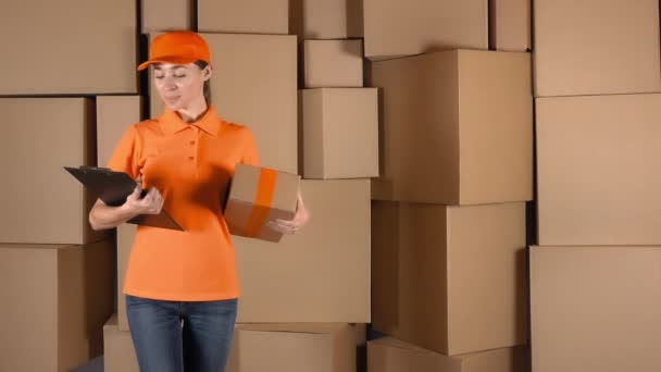 Correio feminino bonito em uniforme laranja entregando um pacote contra caixas de papelão pilhas de fundo. 4K vídeo de estúdio — Vídeo de Stock